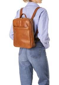 Bruine leren rugzak met een rechthoekige vorm, een voorvak met rits en verstelbare schouderbanden, gedragen door iemand in een blauw gestreept shirt.