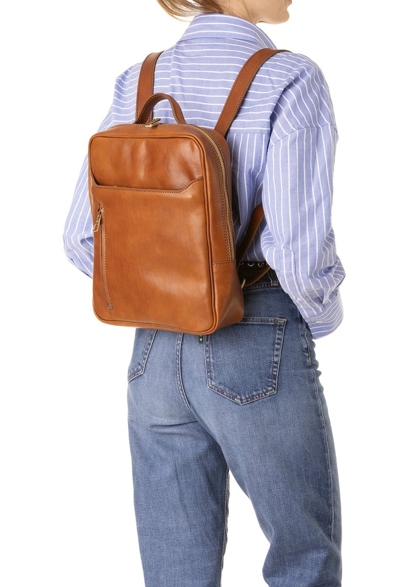 Bruine leren rugzak met een rechthoekige vorm, een voorvak met rits en verstelbare schouderbanden, gedragen door iemand in een blauw gestreept shirt.