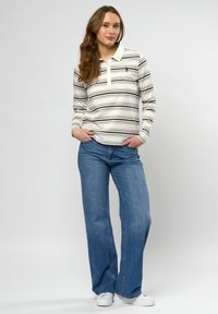 Vrouw met lang haar, gekleed in een witte en zwarte gestreepte lange mouwen polo, blauwe jeans en witte sneakers, staand met handen in haar zakken.