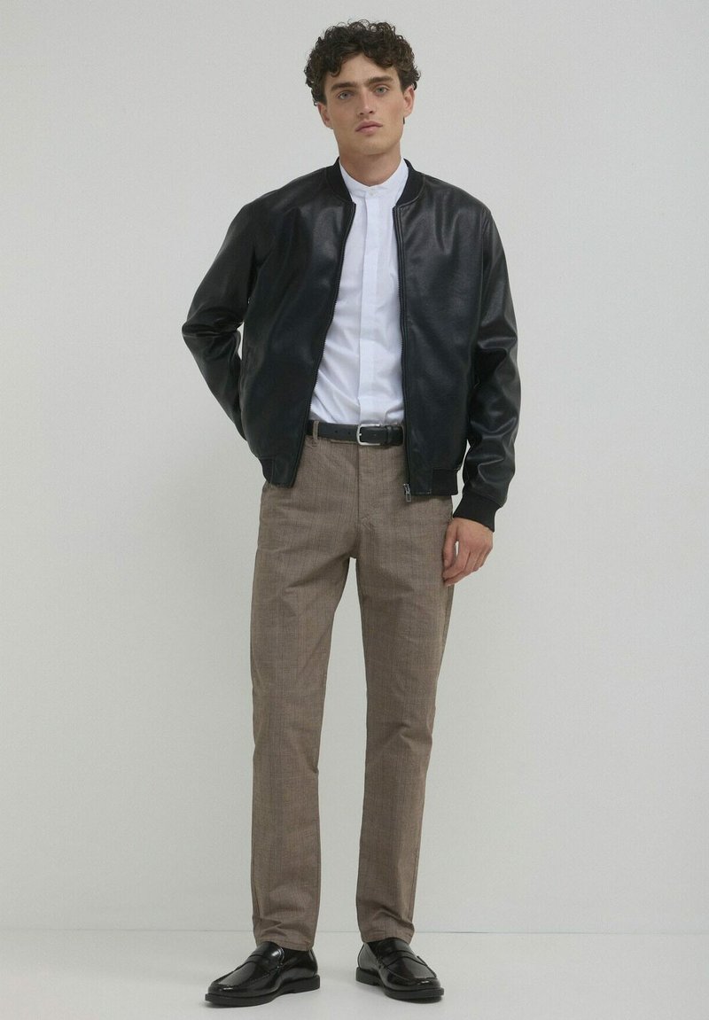Schwarze Leder-Bomberjacke über einem weißen Hemd mit Kragen, kombiniert mit hellbraunen, maßgeschneiderten Hosen und schwarzen Schuhen. Schlichter Hintergrund.