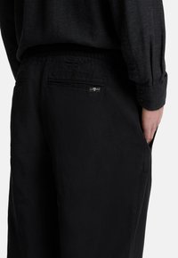 Schwarze Hose mit lockerer Passform, aus leichtem Stoff gefertigt. Verfügt über eine Gesäßtasche mit Nähdetails und einem kleinen Logosticker.