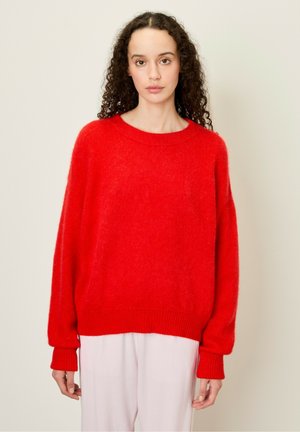 Pull rouge et duveteux en matériau doux avec un col rond. Doté de manches longues et amples et de poignets côtelés. Porté sur un pantalon clair.