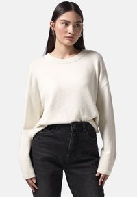 Kremowy, oversize'owy sweter z prążkowanym dekoltem i mankietami, zestawiony z ciemnymi dżinsami o wysokim stanie. Miękka faktura z opadającymi ramionami.