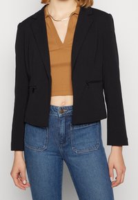Blazer nero cropped con rever a punta, due tasche frontali con zip e vestibilità sartoriale, abbinato a una maglia marrone e jeans blu a vita alta.