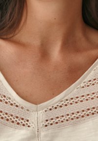 Blusa blanca con escote en V, con detalles de inserciones de encaje en los hombros y una tela lisa y texturizada.