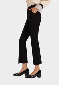 Pantaloni neri su misura con taglio dritto, dotati di tasche laterali e una texture liscia, abbinati a décolleté nere in pelle lucida con dettaglio a fibbia.