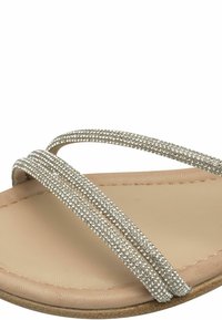 Rihma ja hõbedaste rhinestone'idega kaunistatud kahe metallik hõbedase bändiga cross-sandaal, mis on paigutatud beežile pehmendusega tallale.