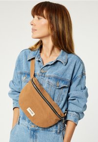 Sac en toile marron en bandoulière, de forme courbée, avec une fermeture éclair noire et une étiquette. La sangle est réglable, posée sur une tenue en denim bleu.