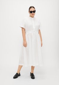Weißes, kurzärmeliges Hemdkleid mit Knopfverschluss und Kordelzug in der Taille. Aus glattem Baumwollstoff, knöchellang und kombiniert mit schwarzen Slippern.