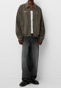 Šedá bavlněná bomber bunda s elastickými manžetami a lemem, obsahující boční kapsy a výšivku, spárovaná s volnými tmavě šedými džíny a černými botami.