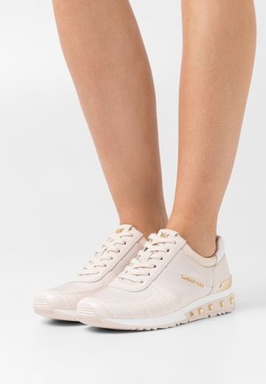 Femme portant des baskets Michael Kors beige clair avec des accents dorés et une semelle cloutée, debout sur une surface blanche.