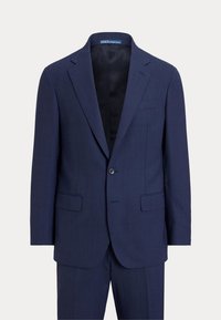 Veste de costume homme bleu marine à un bouton, avec revers crantés et poches à rabat, portée avec un pantalon assorti sur un fond uni.