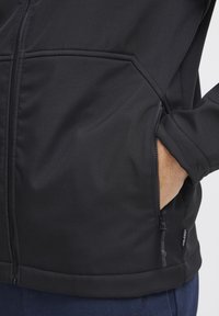 Zwarte zip-up jack met een zachte stof, zijzakken en een nauwsluitende pasvorm met elastische manchetten. Minimale branding nabij de zoom.