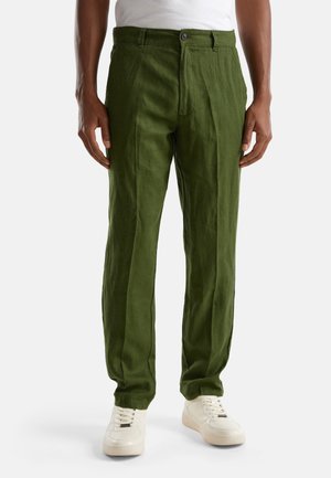 Mann trägt olivgrüne Leinenhose und weiße Sneakers, steht vor einem schlichten weißen Hintergrund.