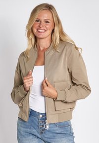 Hellbeige Bomberjacke mit Reißverschluss vorne, gerippten Bündchen und einer Seitentasche, aus einem glatten Stoff. Kombiniert mit einem weißen Oberteil und Jeans.