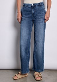 Jean large à taille mi-haute en denim bleu délavé avec une fermeture à bouton, comportant des poches avant et une texture de frottement subtile.