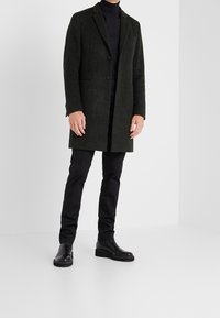 Abrigo de mezcla de lana verde oscuro con corte recto, solapas con muesca, tres botones y bolsillos laterales. Llevado sobre un cuello de tortuga negro y pantalones negros.