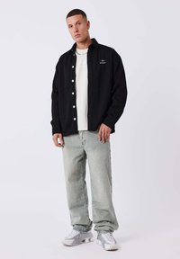 Camisa negra de botones con textura acanalada y botones blancos, sobre una camiseta blanca, combinada con pantalones vaqueros holgados verde claro y zapatillas plateadas.