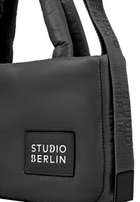 Sort polstret taske med rektangulær form, to korte håndtag og en struktureret rem. Viser et mærke med "STUDIO BERLIN" i hvidt.