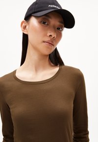 Zwarte baseballpet met "PERMAN" tekst, gedragen met een olijfgroene longsleeve shirt met een ronde hals en een gladde textuur.