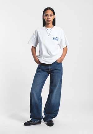 Hvid oversized t-shirt med blå "TACO" tekst, parret med løse blå jeans. Sorte sko med et mønster fuldender outfittet.