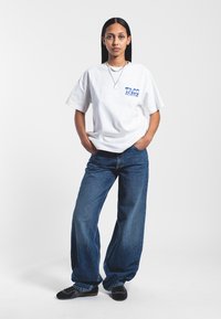 Bílé oversized tričko s modrým nápisem "TACO", kombinované s volnými modrými denimovými džíny. Černé boty s vzorovaným designem dotvářejí outfit.