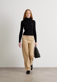 Mustmustritsiga must turtleneck svettari koos väikese logoga, beežid laiad püksid ja must käekott, kandmisega mustade kingadega.