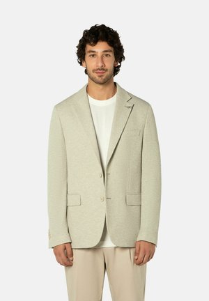Mann mit lockigem dunklem Haar trägt beige strukturierten Blazer über weißem Hemd und hellfarbener Hose, steht vor einfachem Hintergrund.