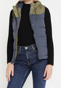 Hooded pufferväst med en färgblockdesign i grön och blå, med dragkedjestängning och snörningsdetaljer. Strukturerat material med vadderad mönstring.