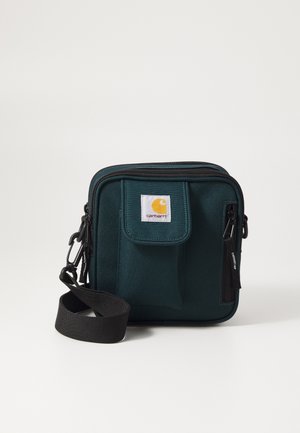 Bolso cuadrado en color teal fabricado con tejido duradero. Cuenta con cremalleras negras, un bolsillo con solapa en la parte frontal y una correa desmontable. Logo de Carhartt exhibido.