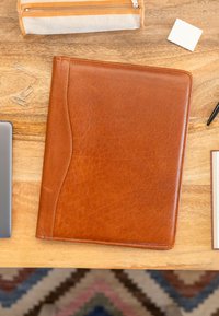 Carpeta de cuero marrón con bordes cosidos y una solapa curvada. Rodeada de un estuche de lápices, un cuaderno y clips de papel sobre una superficie de madera.