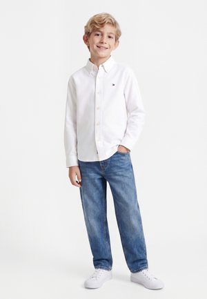 Blond gutt smiler, iført en hvit skjorte med knapper, blå jeans og hvite joggesko, står med en hånd i lommen på hvit bakgrunn.