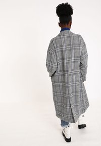 Cappotto lungo a fantasia a quadri in grigio e blu navy, caratterizzato da una linea dritta, maniche ampie e un colletto blu. La vista dal retro mostra l'orlo spaccato e un outfit a strati.