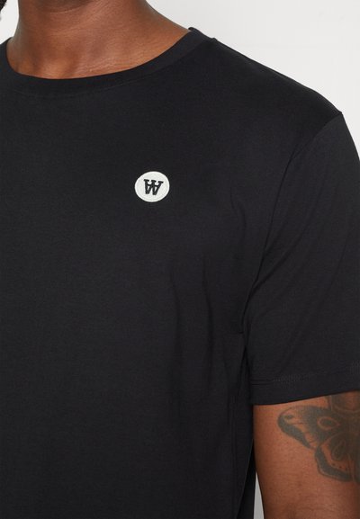 Svart bomulls t-skjorte med ribbestrikket rund hals og en liten hvit sirkulær logo på brystet. Korte ermer, glatt tekstur, tettsittende design.