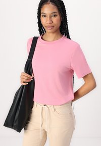 Pinks t-skjorte med korte ermer i et glatt stoff, i kombinasjon med beige stripete bukser. Modellen holder en svart teksturert veske med skulderreim.
