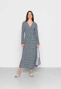 Vestido maxi em azul-marinho com padrões florais brancos, apresenta um design cruzado e mangas longas. Combinado com sandálias de salto bloco em azul claro e uma bolsa.