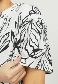 Hvid t-shirt med sort blomster- og bladdesign, med korte ærmer og en afslappet pasform, vist med en hånd der holder kanten.