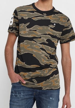 Mand iført en kortærmet t-shirt med camouflageprint og et lille hvidt logo på brystet samt slidte blå jeans.