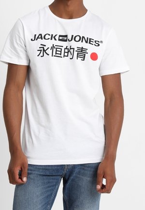 Homme portant un T-shirt blanc Jack & Jones avec texte noir, caractères chinois et un point rouge, associé à un jean bleu.