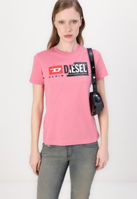 Pink cotton T-shirt with short sleeves features a diagonal seam and a bold black and red logo. Paired with gray jeans and a small black bag.