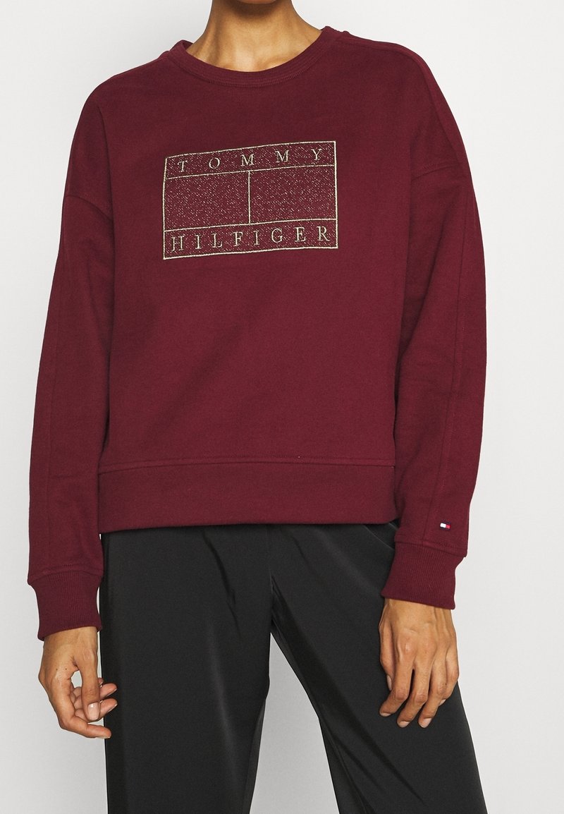 Personne portant un sweat-shirt Tommy Hilfiger bordeaux avec un logo sur la poitrine et un pantalon noir, debout devant un fond uni.