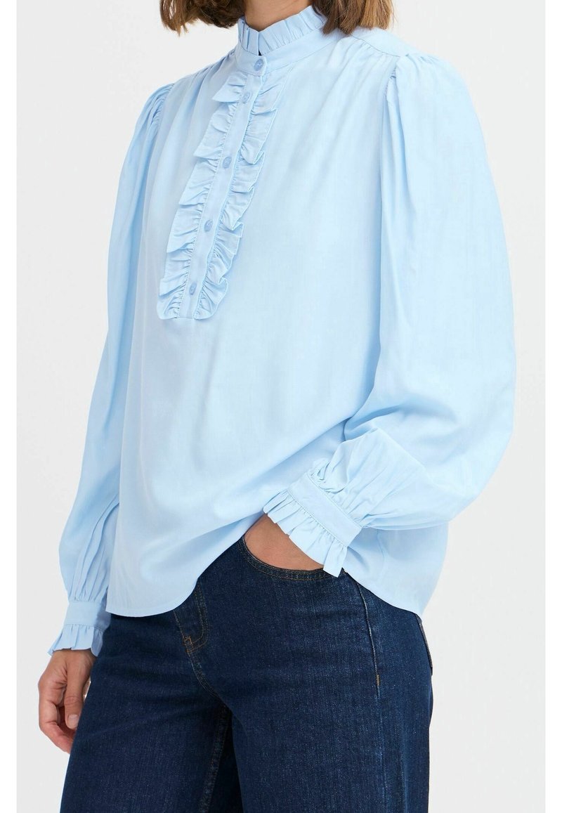 Femme portant un chemisier bleu clair à manches longues avec col montant à volants, patte de boutonnage à l'avant et poignets volantés, associé à un jean foncé.