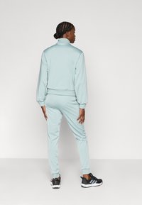 Φωτεινό γαλάζιο σετ φόρμας με λαιμόκοψη ψηλή και μανίκια με ρεβέρ, που περιλαμβάνει παντελόνια στενής γραμμής και μαύρα αθλητικά παπούτσια με λευκές λεπτομέρειες.