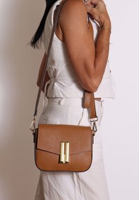 Borsa a tracolla in pelle marrone con texture, dotata di una chiusura in metallo dorato, caratterizzata da una tracolla corta e un design liscio e strutturato.