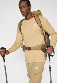 Μπλουζάκι με μακριά μανίκια, παντελόνι που ταιριάζει και πράσινο σακίδιο πλάτης. Εξοπλισμένο με μπαστούνια πεζοπορίας και ζώνη μέσης. Διαθέτει λογότυπα ως διακοσμητικά στοιχεία.