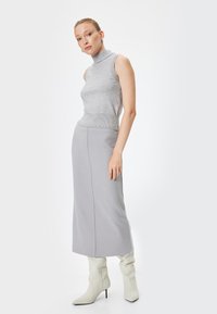 Grijze mouwloze coltrui gecombineerd met een lange grijze rok. Model draagt kniehoogde witte laarzen met spitse neuzen. Gladde texturen, minimalistisch ontwerp.