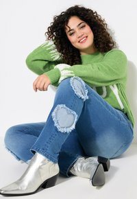 Suéter verde de punto con letras blancas, jeans azules desgastados con parches en forma de corazón y botas plateadas hasta el tobillo con tacón cuadrado.