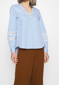 Mujer vistiendo una blusa de rayas azul y blanca con un escote en V y detalles de encaje en las mangas, junto con pantalones de pierna ancha marrones, frente a un fondo blanco.