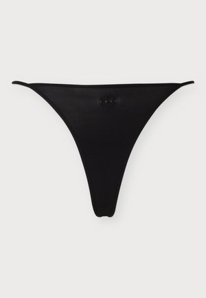 Mujer descalza vistiendo un bikini negro con tirantes delgados sobre un fondo liso y claro, mirando al frente con una ligera sonrisa.