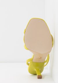 Sandale à talons aiguilles jaunes avec un supérieur en tissu doux, semelle lisse et brillante, talon aiguille étroit, et un petit accent en bois à la base du talon.